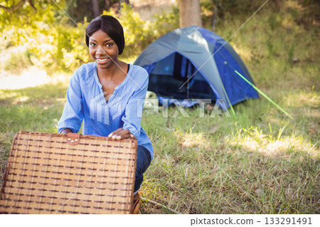 African American woman kneeling holding picnic basket lid open in forest clearing, copy space 133291491