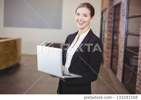 Female professional holding silver laptop while standing at office lobby wearing black blazer Female professional holding silver laptop while standing at office lobby wearing black blazer 133291508