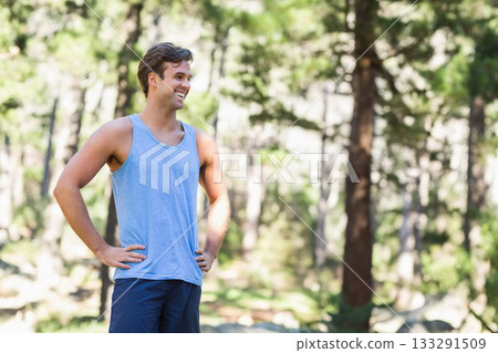 Man standing with hands on hips smiling in pine forest clearing among rocks under dappled sunlight Man standing with hands on hips smiling in pine forest clearing among rocks under dappled sunlight 133291509