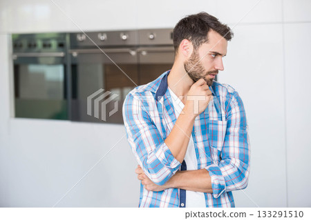 Man in 30s in plaid shirt resting hand on chin in kitchen beside ovens, copy space 133291510