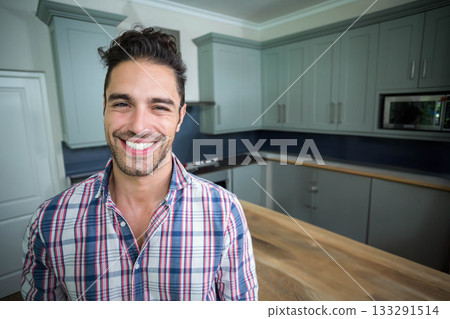 Smiling man standing in kitchen beside wooden island, cabinets and dark backsplash, copy space Smiling man standing in kitchen beside wooden island, cabinets and dark backsplash, copy space 133291514