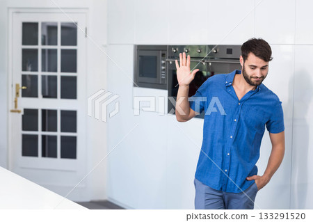 Man in late twenties standing in kitchen by integrated appliances and glass door waving, copy space Man in late twenties standing in kitchen by integrated appliances and glass door waving, copy space 133291520