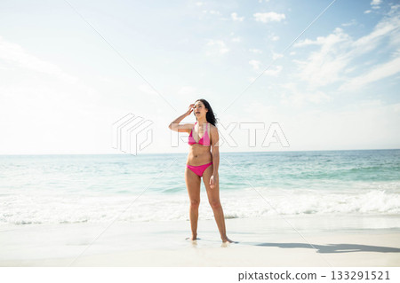 Woman standing barefoot at beach edge wearing pink bikini, shielding eyes and gazing toward horizon 133291521