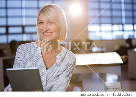 Businesswoman smiling while holding tablet at office desk in patterned blouse with conference table 133291525