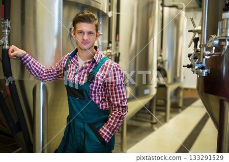 Male brewery worker in overalls standing near fermentation tanks at brewery with gauges, copy space Male brewery worker in overalls standing near fermentation tanks at brewery with gauges, copy space 133291529