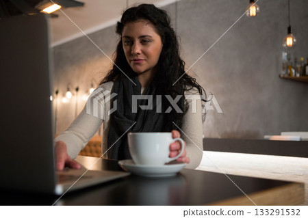 Woman sitting at wooden cafe counter typing on laptop while holding ceramic coffee cup on saucer 133291532