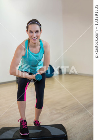 Mid adult woman holding dumbbell and leaning on step platform in fitness studio with exercise ball Mid adult woman holding dumbbell and leaning on step platform in fitness studio with exercise ball 133291535