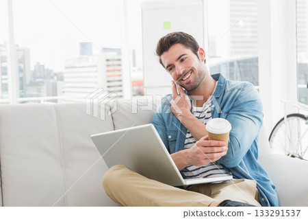 Man in 30s calling on smartphone and typing on laptop at office lounge, bike leaning nearby Man in 30s calling on smartphone and typing on laptop at office lounge, bike leaning nearby 133291537