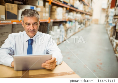 Mature man wearing business attire leaning on box in storage aisle holding tablet, copy space 133291541