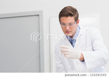 Male scientist examining clear test tube in research lab wearing lab coat and goggles, copy space 133291543