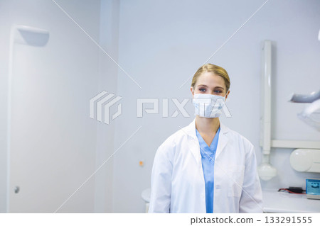 Female medical professional standing in exam room wearing scrubs under lab coat and surgical mask Female medical professional standing in exam room wearing scrubs under lab coat and surgical mask 133291555