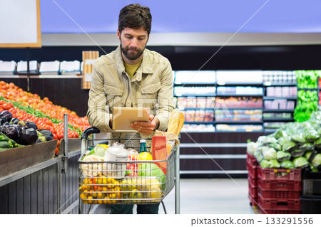 Male shopper consulting tablet while pushing cart by produce bins in supermarket aisle Male shopper consulting tablet while pushing cart by produce bins in supermarket aisle 133291556