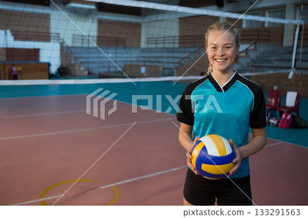 Teenage girl holding volleyball on gym court with net, red chairs and sports bags, copy space 133291563