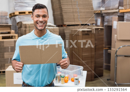 Male warehouse worker holding blank cardboard sign in warehouse amid flattened boxes and container 133291564