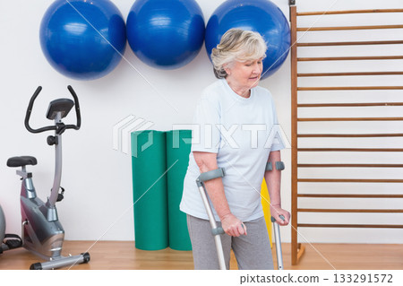 Senior White woman using forearm crutches balancing in rehab gym with balls and bike, copy space 133291572