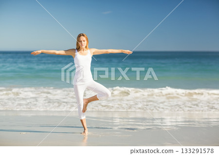 female practicing yoga tree pose at sandy shoreline wearing fitted tank top and cropped leggings 133291578