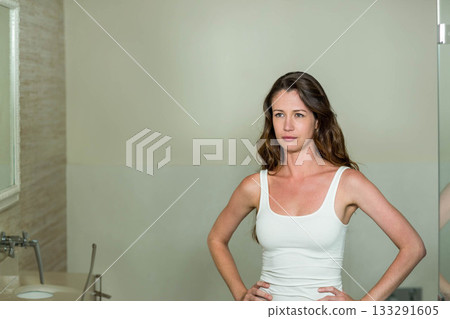 Woman standing hands on hips by sink facing mirror wearing tank top in bathroom, copy space Woman standing hands on hips by sink facing mirror wearing tank top in bathroom, copy space 133291605