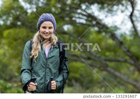 Woman hiking on wooded trail wearing windbreaker and beanie carrying backpack and trekking poles 133291607