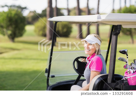 Female golfer gripping steering wheel while sitting in golf cart on sunlit fairway with golf bag Female golfer gripping steering wheel while sitting in golf cart on sunlit fairway with golf bag 133291608