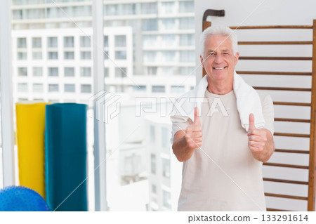 Senior man standing in fitness studio giving thumbs up with towel draped, stall bars, copy space Senior man standing in fitness studio giving thumbs up with towel draped, stall bars, copy space 133291614