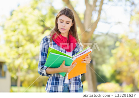 Female student holding two open notebooks standing in sunny park wearing plaid shirt and red scarf 133291627