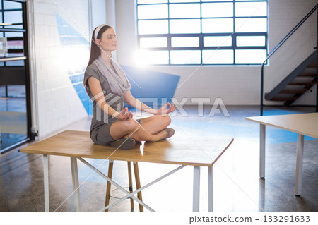 Businesswoman wearing gray dress sitting cross-legged on table in office catching grid window flare 133291633