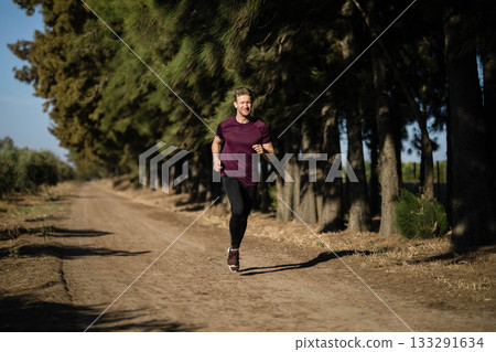 Male jogger running along dirt trail through pine forest wearing maroon athletic shirt Male jogger running along dirt trail through pine forest wearing maroon athletic shirt 133291634
