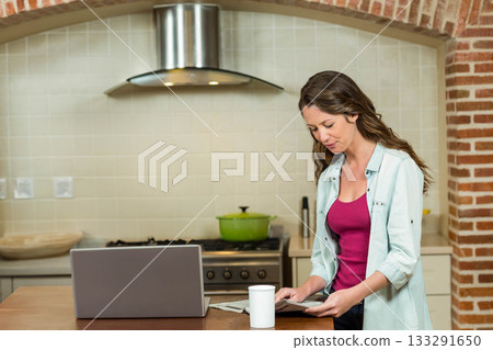 Woman reading tablet while cooking on kitchen island with laptop coffee mug newspaper, copy space 133291650