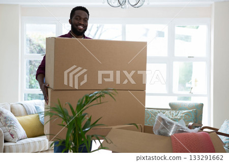 Smiling African American man carrying cardboard boxes in living room with bubble wrap 133291662