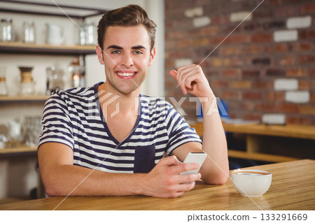 Man smiling while holding smartphone at coffee shop table with ceramic mug near shelves, brick wall 133291669
