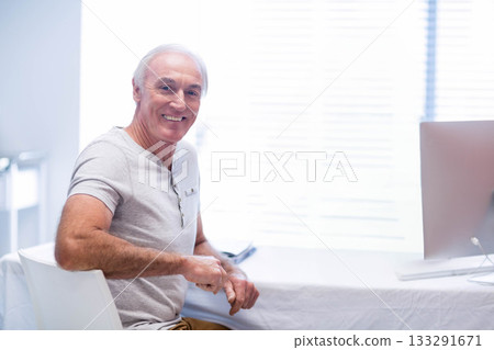 Smiling senior man working on desktop computer in home office with smartphone and window blinds Smiling senior man working on desktop computer in home office with smartphone and window blinds 133291671