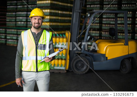 Male warehouse worker wearing helmet, vest holding clipboard near pallets and forklift, copy space 133291673