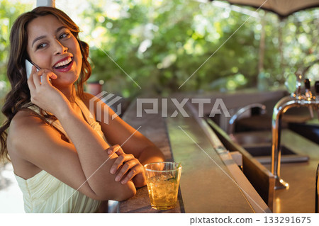 Woman leaning on bar counter at cafe talking on smartphone while sipping iced drink, copy space 133291675