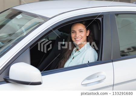 Female driver sitting inside white sedan beside stucco wall, showing steering wheel and side mirror 133291680