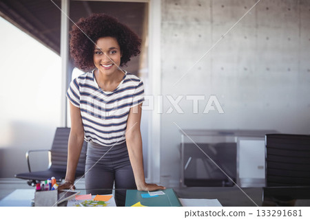 African American woman leaning on glass-top desk in office with sticky notes, pen holder, smiling 133291681