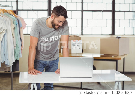 Man volunteering leaning over folding table typing on laptop in warehouse beside rack of shirts 133291689