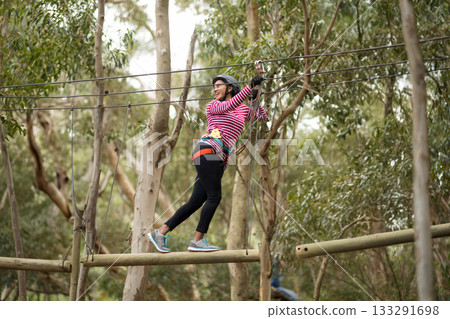 African American woman wearing harness balancing on beam holding overhead cable in ropes course 133291698