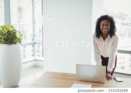 African American woman leaning over desk in modern office smiling with laptop and plant, copy space 133291699