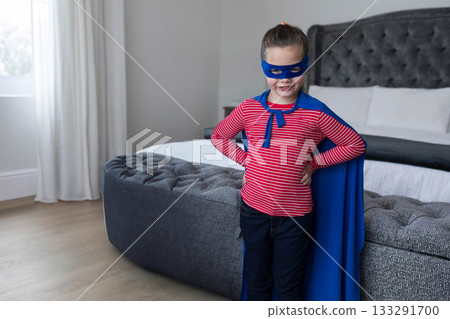 Child girl wearing blue cape, mask, striking pose beside bench at foot of bed, copy space 133291700