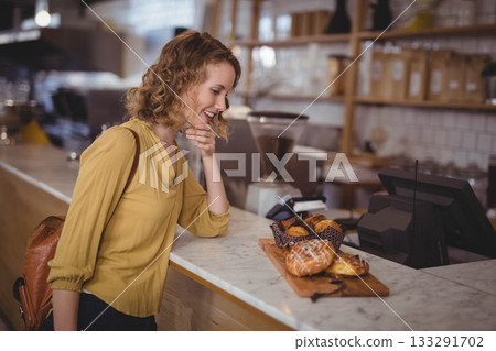 Woman in her 20s leaning toward wooden pastry board at countertop inside cafe, selecting croissant 133291702