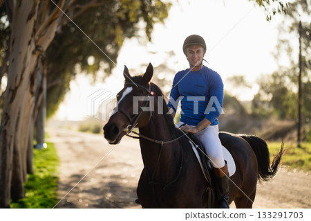 Man riding horse on sunlit trail flanked by eucalyptus trees wearing helmet and riding boots Man riding horse on sunlit trail flanked by eucalyptus trees wearing helmet and riding boots 133291703