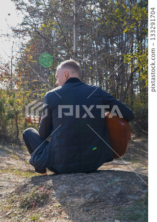 Beautiful blonde man playing acoustic guitar and singing relaxing enjoying outdoors on sunny autumn day. Artist learning and practicing playing musical instrument. Fall leaves autumnal 133291704