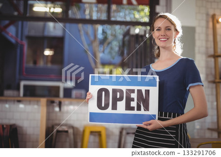 Woman in striped apron holding up blue-and-white open sign inside café at counter, copy space 133291706