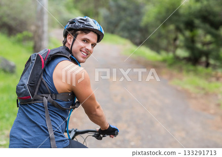 Male cyclist wearing helmet smiling, looking back and riding mountain bike on gravel forest trail Male cyclist wearing helmet smiling, looking back and riding mountain bike on gravel forest trail 133291713