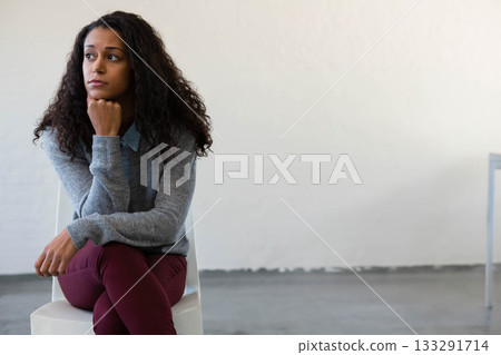 Woman sitting pensively on white chair in studio wearing grey sweater with table leg, copy space Woman sitting pensively on white chair in studio wearing grey sweater with table leg, copy space 133291714