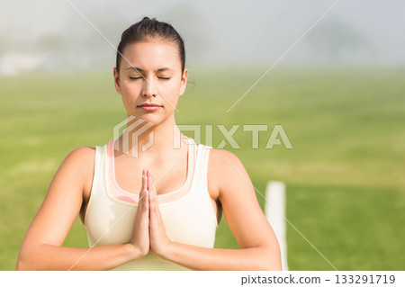 Woman standing in grassy field wearing light-colored sports top pressing hands together in prayer 133291719