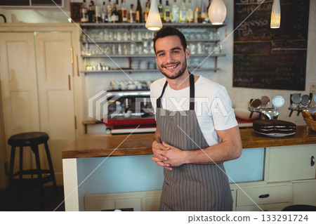 Male cafe worker wearing striped apron standing and smiling behind counter with espresso machine 133291724