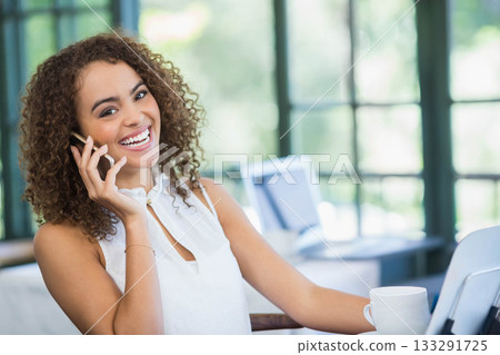 African American woman holding smartphone, smiling at desk in bright office with laptop, coffee mug 133291725