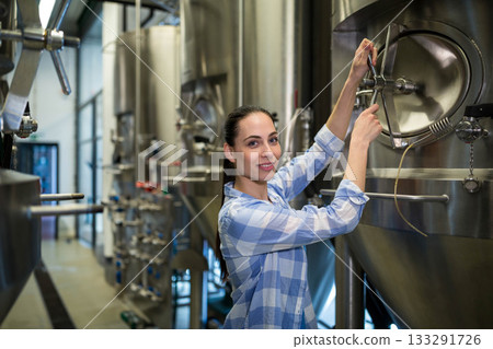 Female brewer adjusting latch on stainless steel fermentation tank amid valves in brewery facility 133291726