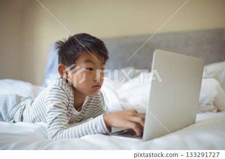 Asian child boy lying on bed using silver laptop on rumpled white bedding in bedroom Asian child boy lying on bed using silver laptop on rumpled white bedding in bedroom 133291727